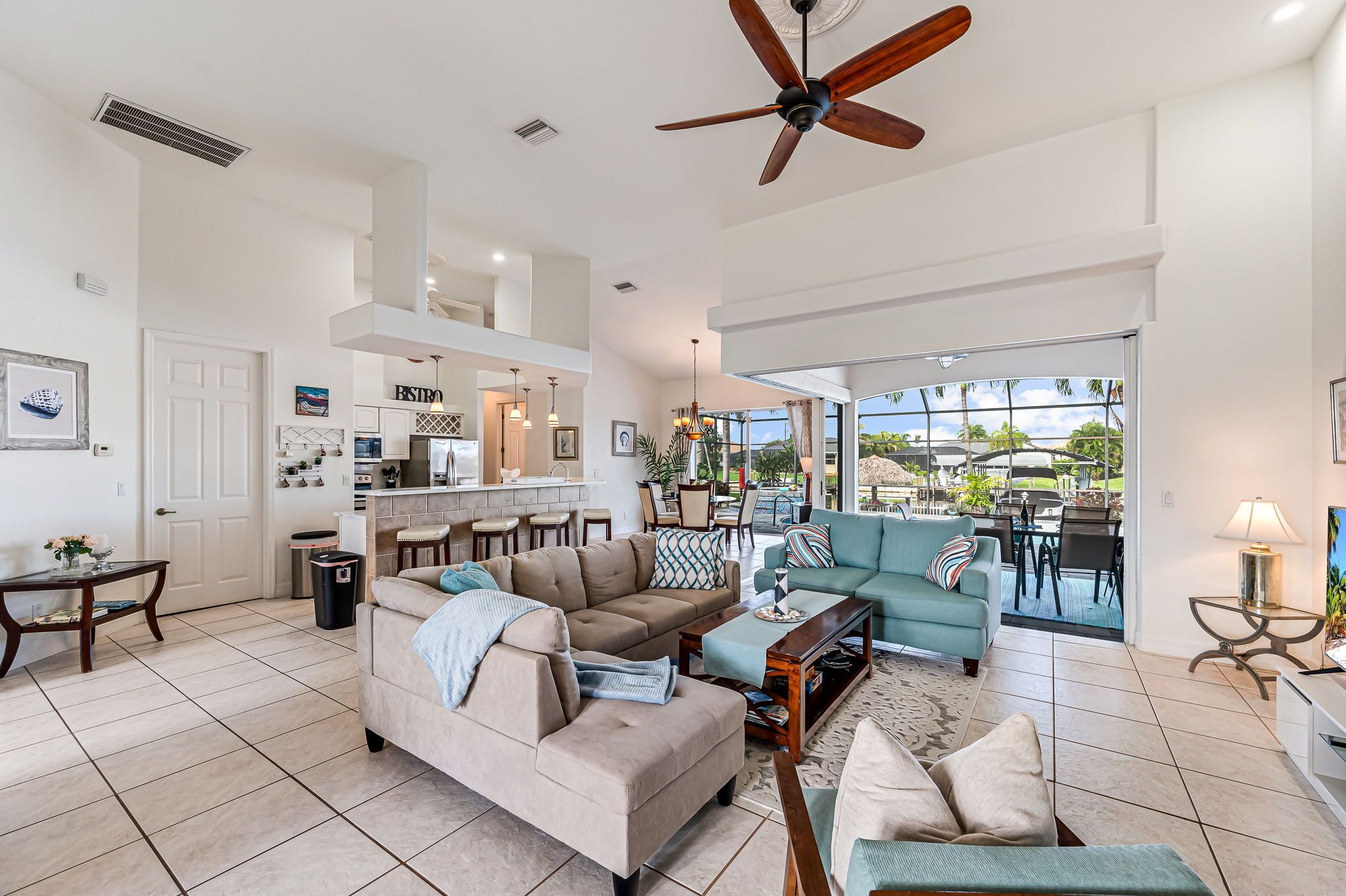 A spacious living area with a sofa, coffee table, and a dining area with chairs. A ceiling fan is visible, and there is a view of the outdoors through large windows.