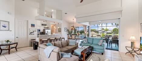 A spacious living area with a sofa, coffee table, and a dining area with chairs. A ceiling fan is visible, and there is a view of the outdoors through large windows.