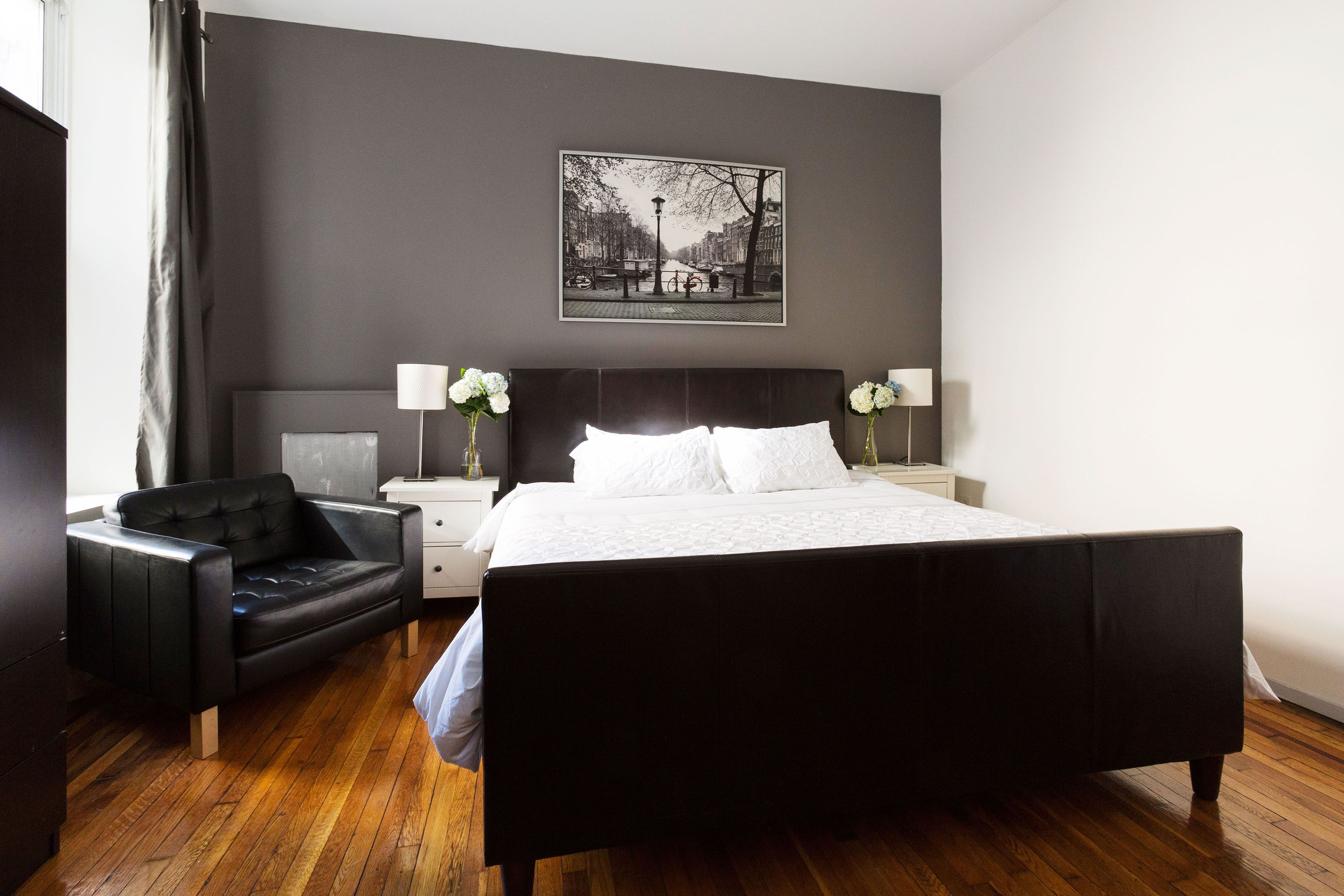 A modern hotel room with a large bed, a black leather chair, a nightstand with a lamp, and a framed cityscape picture on the wall.