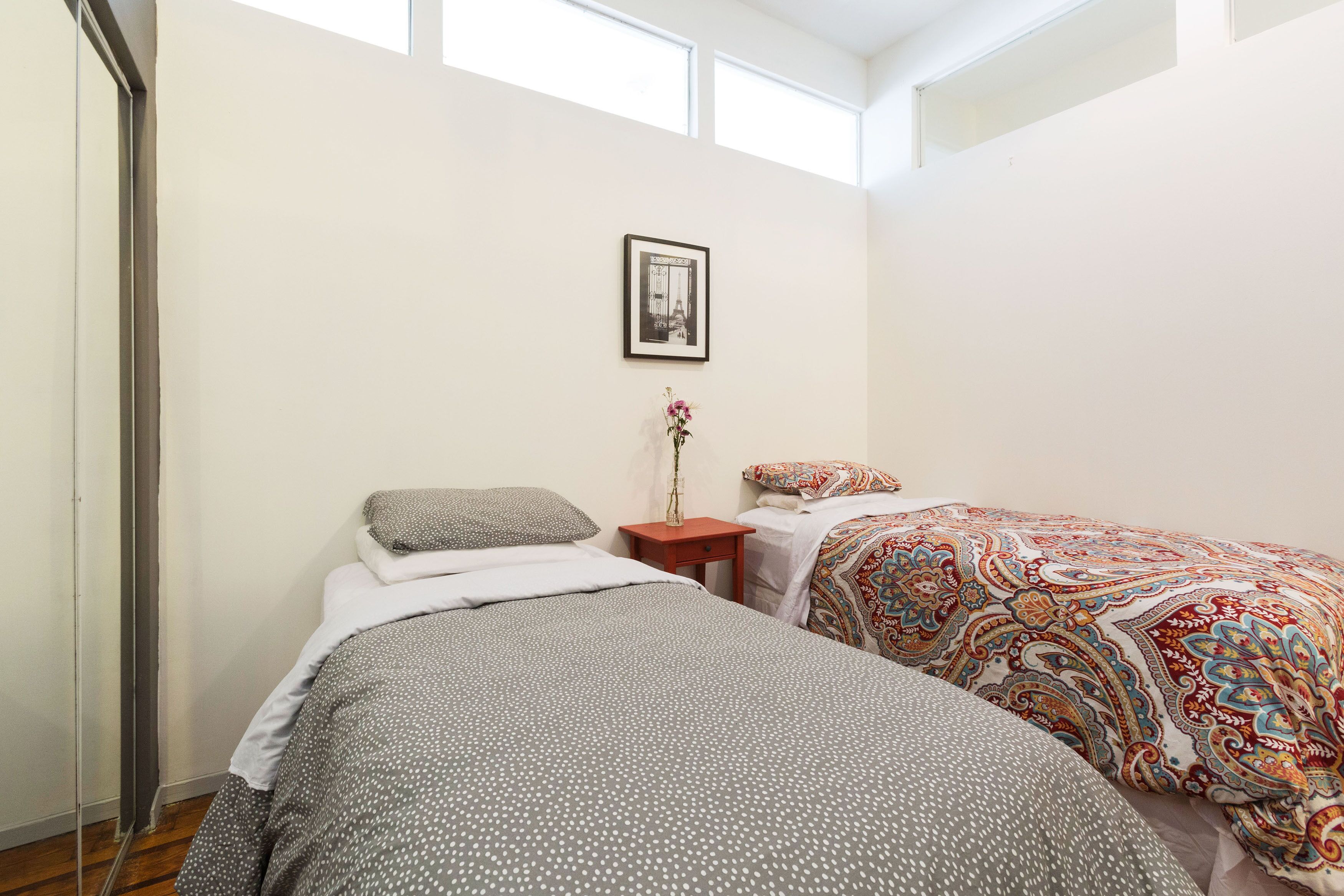 A bedroom with two beds, a small wooden nightstand, a framed picture on the wall, and a window providing natural light.