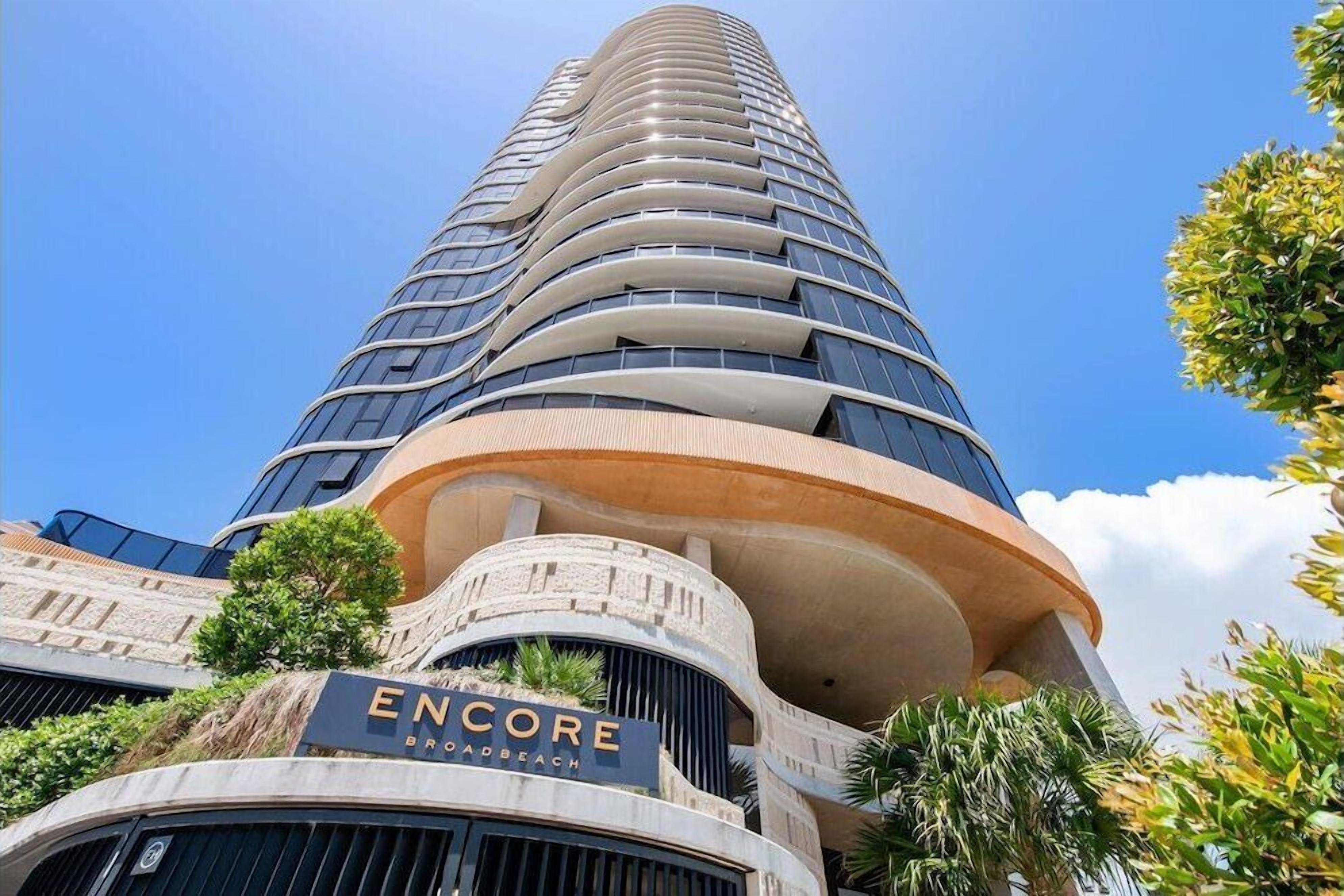 A tall, modern hotel with a curved facade and a sign reading "ENCORE BROADBEACH".