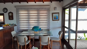 A dining area with a glass table, white chairs, a wooden ceiling, a window with blinds, and a framed picture on the wall.