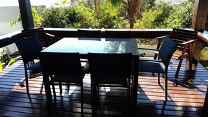 A wooden deck with a glass table and four chairs, surrounded by lush greenery.