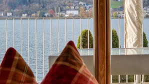 Une vue depuis une fenêtre, avec un coussin à carreaux, donnant sur un lac, des maisons et des collines.