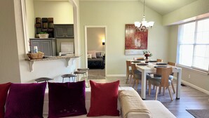 Un salon moderne avec un canapé, un coin repas comprenant une table et des chaises, une cuisine équipée de placards et une grande fenêtre laissant entrer la lumière naturelle.