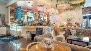 En hyggelig café med en buet disk, siddeområde med sofaer og et rundt bord, samt en væg med hængende planter og et skilt, der står 'The Palm Coffee'.