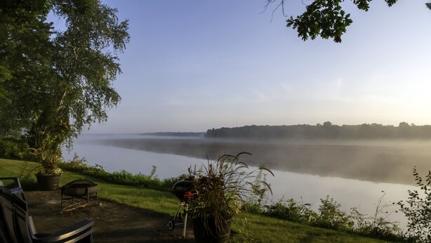 A lakeside view with a bench and a grill, surrounded by greenery and a clear sky.