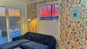 A cozy living room with a blue sofa, a colorful coffee table, and a view of a sunset through a window.