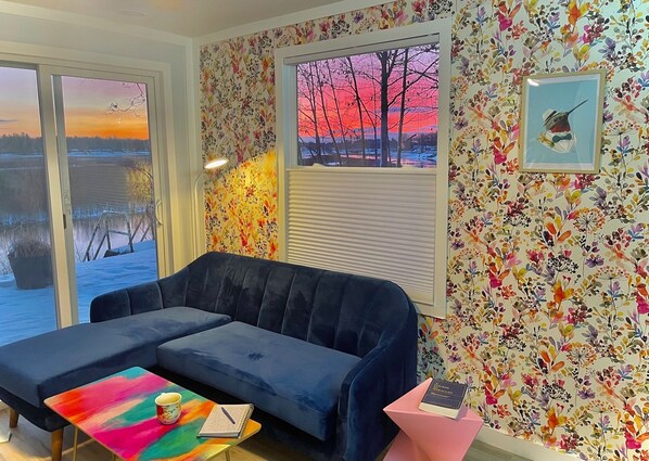 Una sala de estar acogedora con un sofá azul, una mesa de centro colorida y vistas a un atardecer a través de una ventana.
