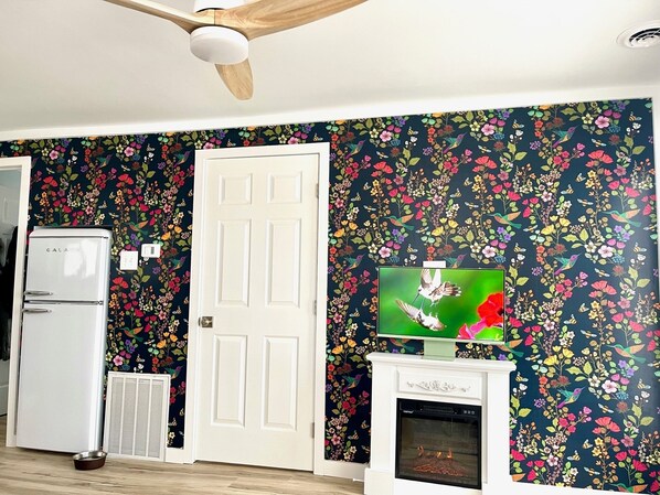 A room with a white door, a white fireplace, a television on a stand, and a ceiling fan.