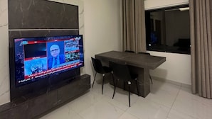 A modern living room with a flat-screen TV mounted on the wall, a dining table with chairs, and an air conditioning unit.