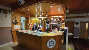 Uma área de bar em madeira com balcão, diversos frascos e uma placa com a inscrição "THE HELM".