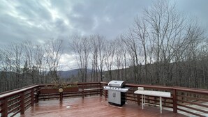 A wooden deck with a barbecue grill and a table, surrounded by trees and a cloudy sky. - Ski Retreat Townhome in Plymouth, VT (Plymouth)