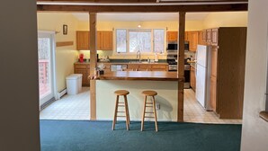 A kitchen with wooden cabinets, a central island with stools, a refrigerator, and a window with a view of trees. - Ski Retreat Townhome in Plymouth, VT (Plymouth)