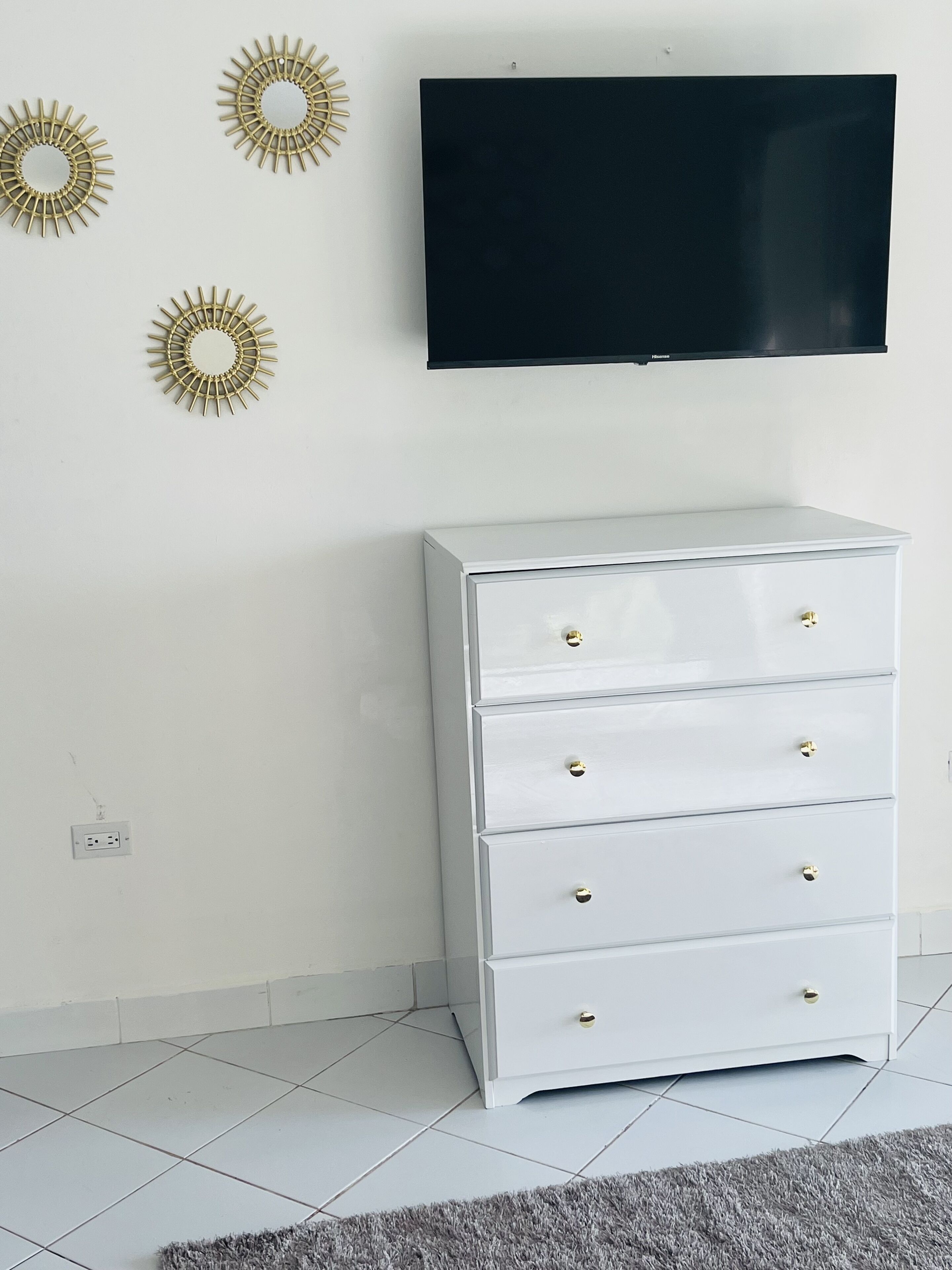 A white dresser with gold knobs, a flat-screen TV mounted on the wall, and three gold sunburst wall decorations.