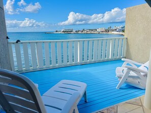 Ein Balkon mit weißen Plastikstühlen und blauem Boden, mit Blick auf Strand und Stadtlandschaft.