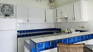 A kitchen with white cabinets, a blue-tiled backsplash, a white countertop, a chandelier, and a wicker chair. - Nice and cozy ocean front studio with private terrace. Short walk to beaches. (Beacon Hill)