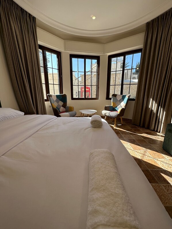 A hotel room with a large bed, two armchairs, a small table, and a view of a building through large windows.
