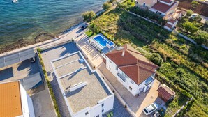 An aerial view of a coastal property with a swimming pool, a tennis court, and a small garden.