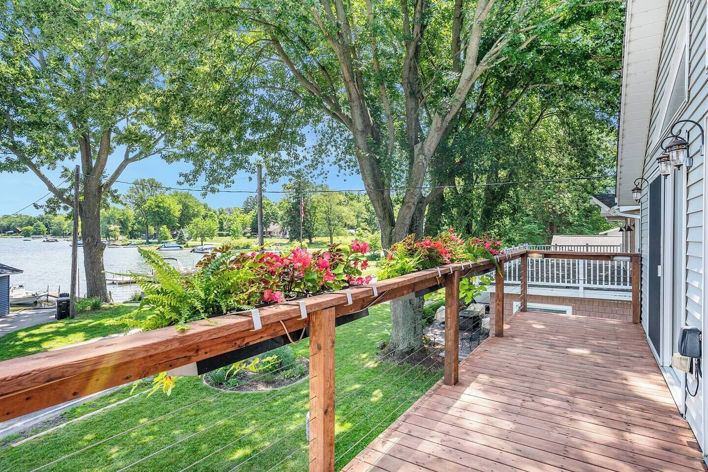 A wooden deck with a railing, overlooking a lake with boats, surrounded by lush greenery and flowering plants.