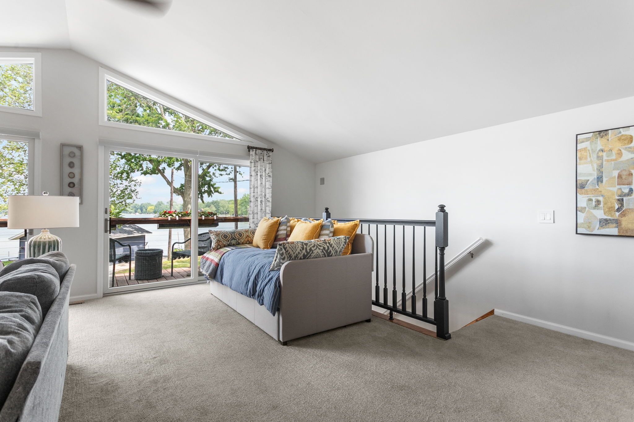 Un dormitorio moderno con un ventanal grande, una cama con almohadones estampados, un sofá gris y una escalera.