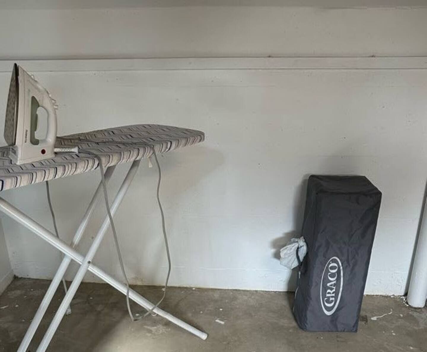 An ironing board with an iron and a Graco bag against a white wall.