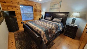 A bedroom with a bed, wooden walls, a ceiling fan, a window, a TV, a dresser, and a lamp.