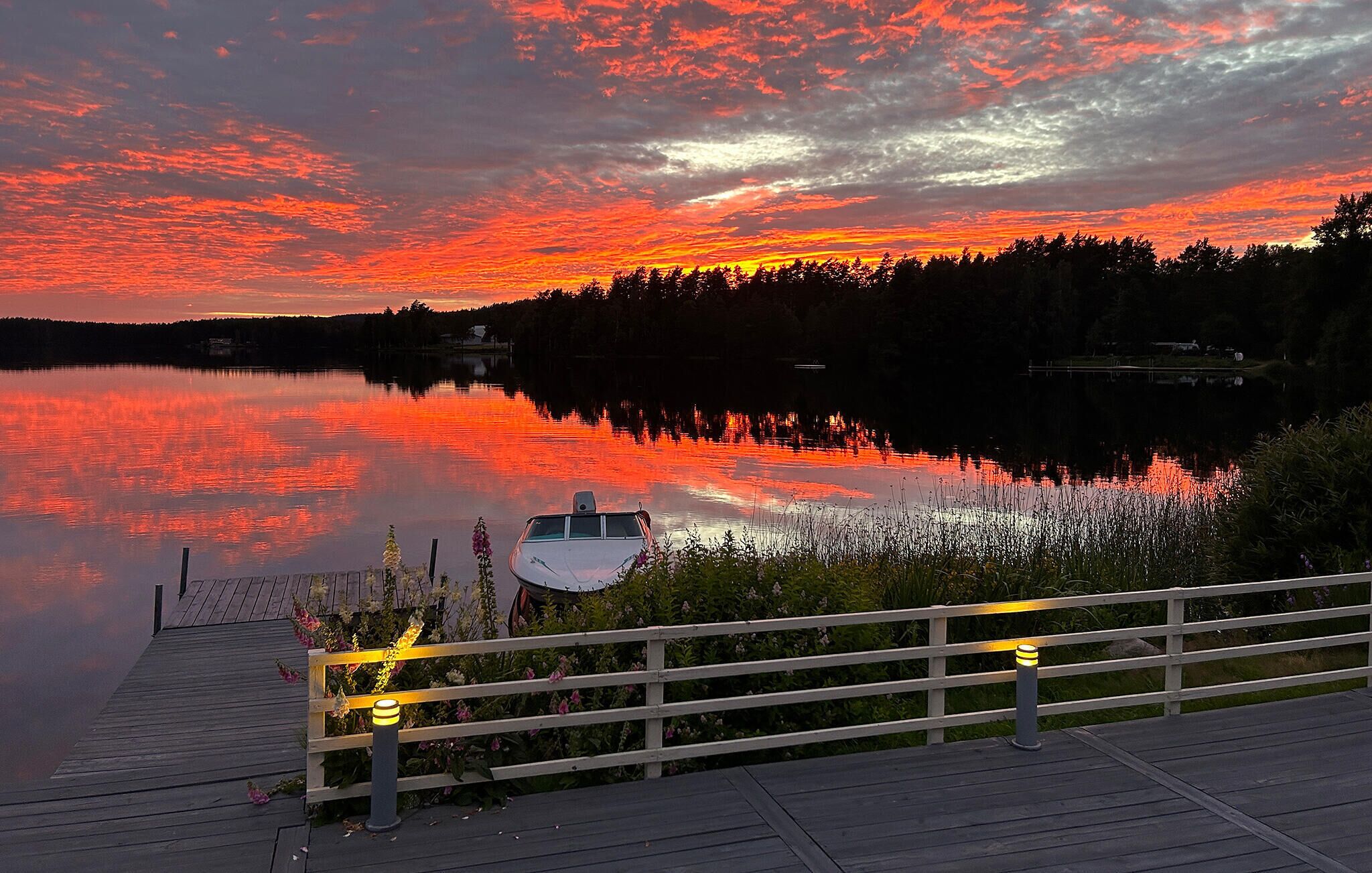Un molo di legno con ringhiere e una barca, affacciato su un lago calmo con il riflesso del tramonto.