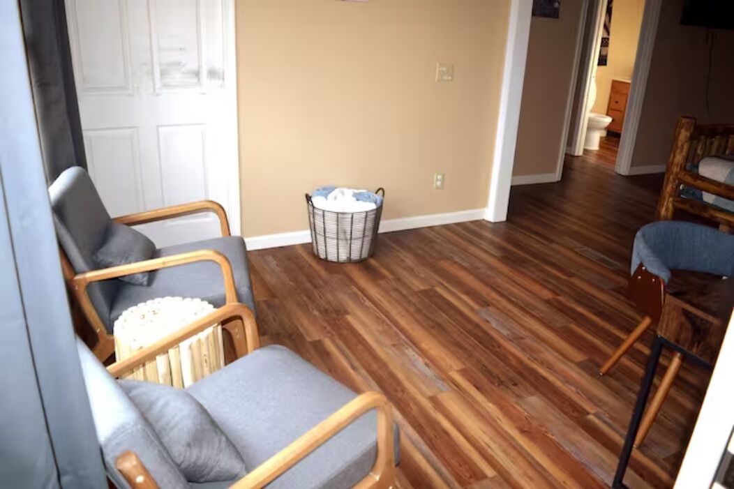 A room with wooden flooring, a white door, and two wicker chairs with cushions.