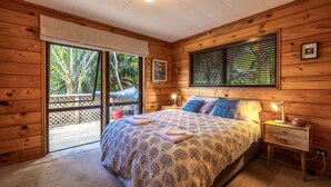 Une chambre dans une cabane en bois, comprenant un lit, une table de chevet, une fenêtre avec des stores et une porte donnant sur un espace extérieur.