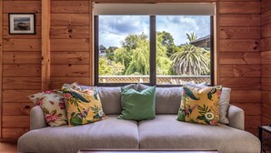 L'intérieur d'un chalet en bois, avec un canapé beige, des coussins à motifs floraux et une vue sur un paysage tropical, visible à travers une grande fenêtre.