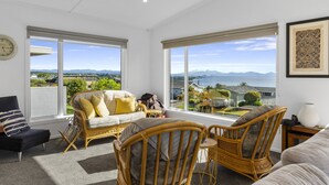 Un salon spacieux doté d’une grande fenêtre offrant une vue sur la mer et les montagnes. On y trouve un canapé, deux fauteuils en osier et un fauteuil noir.