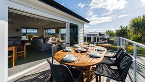 Un espace repas extérieur avec une table en bois et des chaises noires, un canapé et une cuisine visible à travers une porte coulissante ouverte.