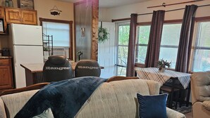 A living room with a beige sofa, two pillows, a ceiling fan, a window with curtains, and a kitchen area with a refrigerator and cabinets.