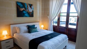 Una cama bien hecha con sábanas blancas y un edredón negro, una mesita de noche con lámpara, y un ventanal con cortinas.