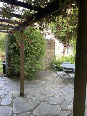 Een terras van stenen met een pergola vol rode bloemen, een stenen muur, en een tafel met stoelen.