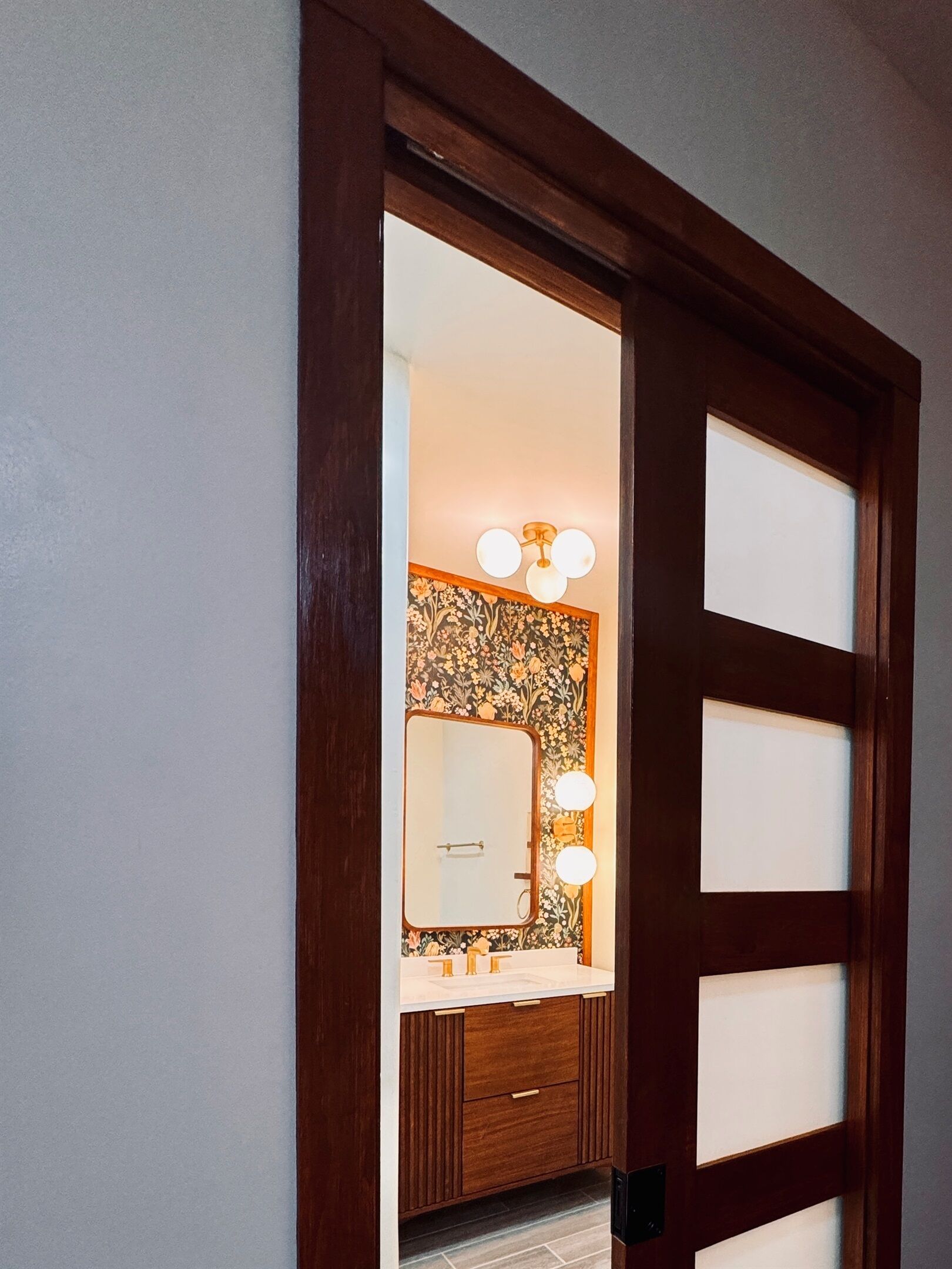 A wooden door with frosted glass panels, leading to a bathroom with a mirror, two sinks, and a floral-patterned wall.