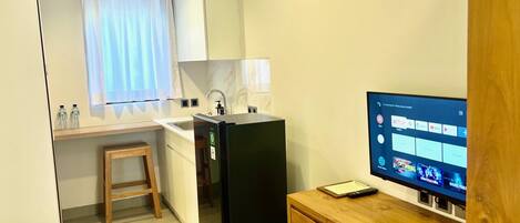 Quarto de hotel compacto com cozinha compacta, uma escrivaninha de madeira, um frigorífico preto, televisão e janela com cortinas.