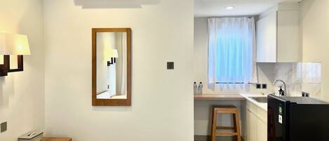 Habitación de hotel con cama, mesita de noche, lámpara, equipo de aire acondicionado, espejo y una kitchenette con refrigerador, lavamanos y encimera.