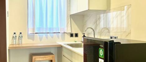 Cocina pequeña y funcional con refrigerador integrado, un taburete de madera, una ventana con cortinas traslúcidas y salpicadero de azulejos.