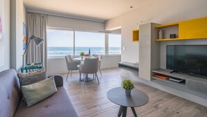 A modern living room with a sofa, a dining table with chairs, a small round table with a plant, a TV, and a view of the ocean.