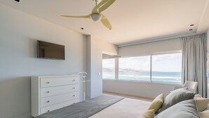 A bedroom with a bed, a ceiling fan, a dresser, a TV, and a large window with a view of the ocean.