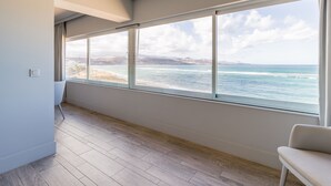 A room with large windows overlooking a beach, wooden flooring, and a white chair.