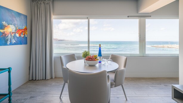 A modern room with a large window overlooking the ocean, a round table with chairs, and a painting on the wall.