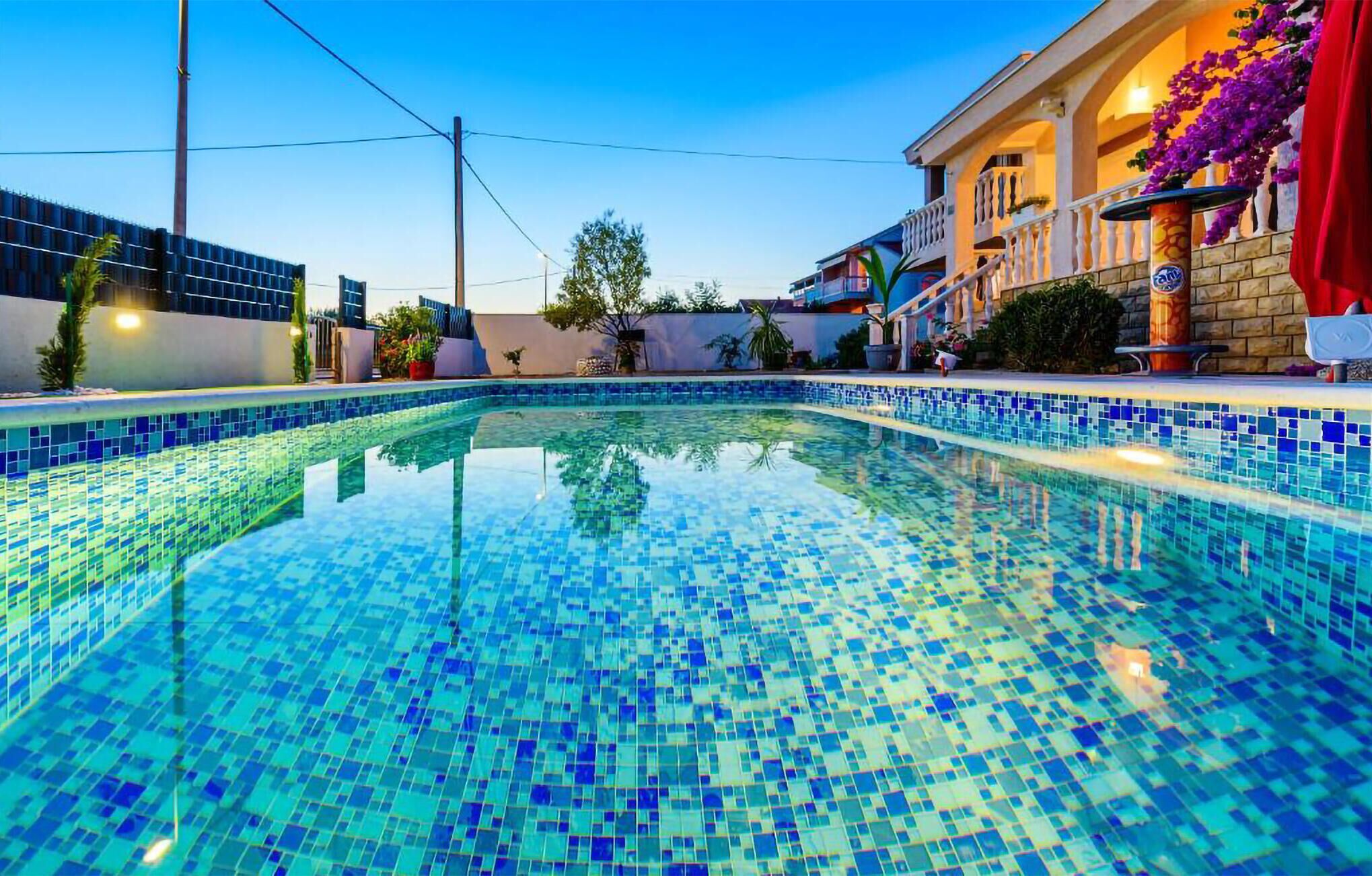 Una piscina con mosaicos azules, rodeada por un edificio con balcón y plantas en macetas.