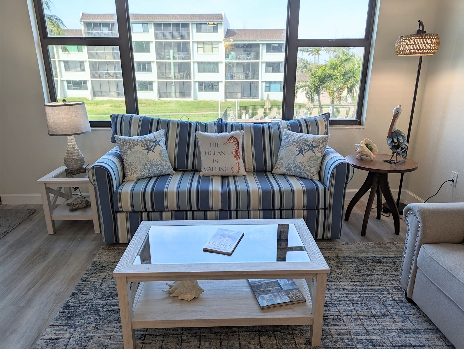 A living room with a striped sofa, a glass coffee table, and a view of a building through large windows.