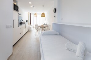 Ein modernes, minimalistisches Hotelzimmer mit einem Einzelbett, einer kleinen Küchenzeile und einem Essbereich mit Tisch und Stühlen.