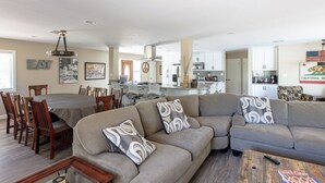 Een ruime woonkamer met een hoekbank, een eettafel en stoelen, een keuken op de achtergrond en een plafondventilator.