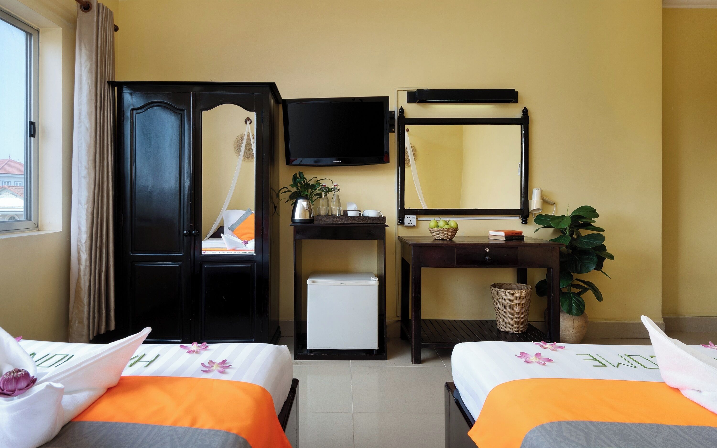 A hotel room with two beds, a TV, a small desk with a mirror, a refrigerator, and a window with curtains.