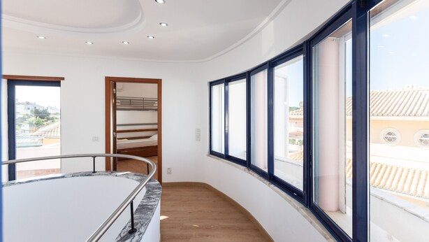 A modern interior with a curved wooden floor, a marble-topped table, and large windows offering a view of the cityscape. - FLH Caparica Town House with Pool (Charneca de Caparica)